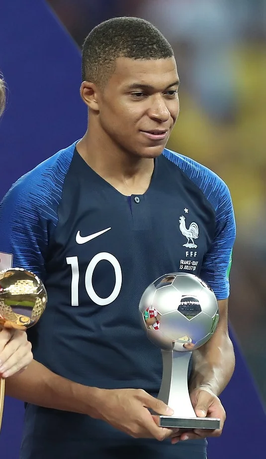 Kylian Mbappé in France's number 10 shirt holding the Best Young Player trophy after the 2018 World Cup final — he captains France into World Cup 2026