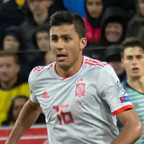 Rodri in Spain's 2026 World Cup home kit — Adidas's deep red shirt with navy sleeves, red-yellow-red shoulder stripes echoing the Spanish flag, thin yellow pinstripes and the word 'España' printed inside the collar, paired with navy blue shorts and socks, released on November 6, 2025 as the senior home jersey La Roja will wear in defence of Euro 2024 at the World Cup in the United States, Canada and Mexico